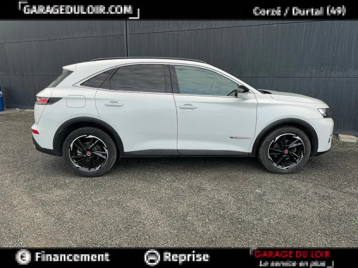 Vue de profil côté gauche d'un DS7 Crossback blanc avec jantes noires et vitres teintées, sur fond gris urbain.