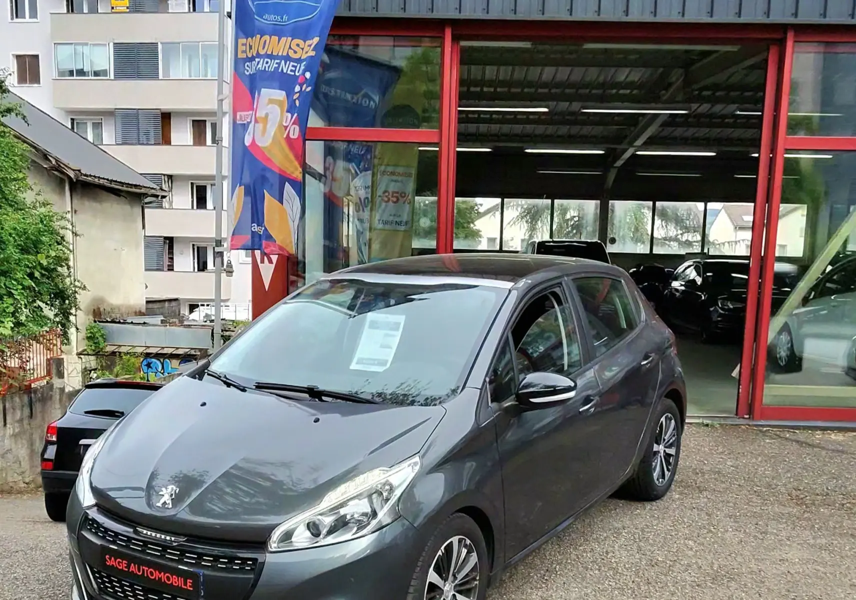 Peugeot 208 gris foncé métal vue 3/4 avant droit devant un garage avec enseigne AD Expert.