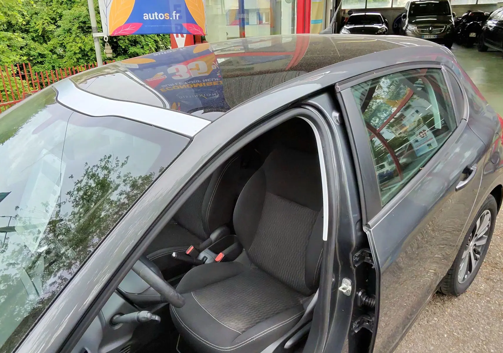Vue côté gauche avant d'une Peugeot 208 gris foncé métal avec porte ouverte, montrant le siège conducteur en tissu noir.