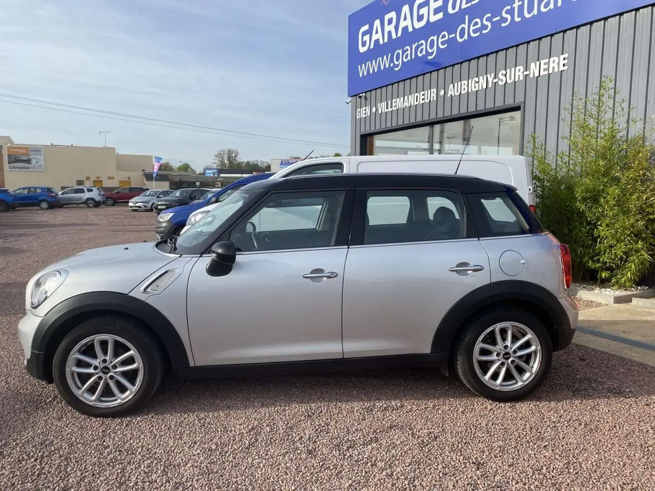 Profil côté gauche d'une MINI Countryman gris clair avec toit noir, garée devant un garage sur un sol gravillonné.