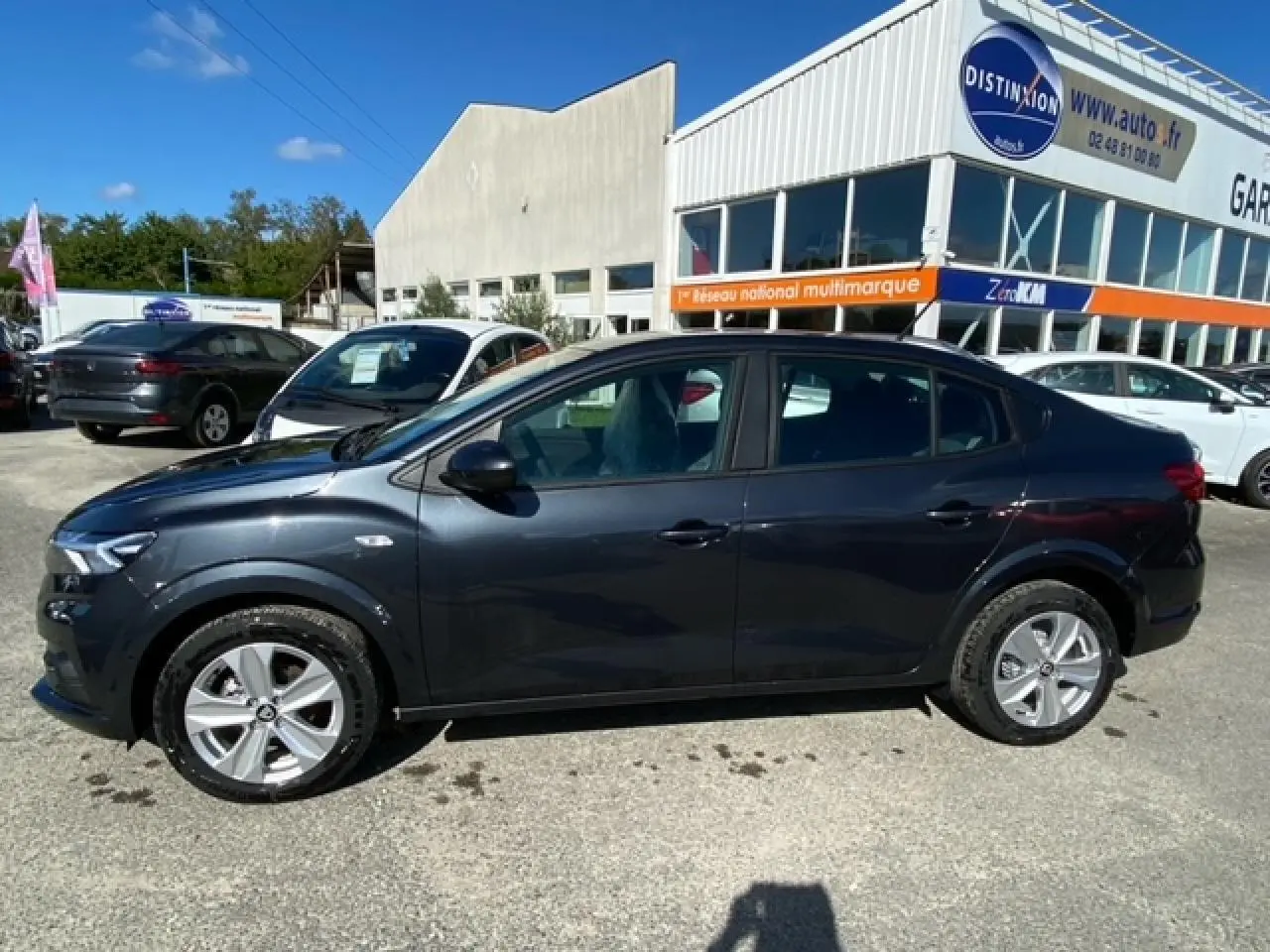 Profil côté gauche d'une Dacia Logan gris foncé 2022 garée devant un garage sous un ciel bleu.