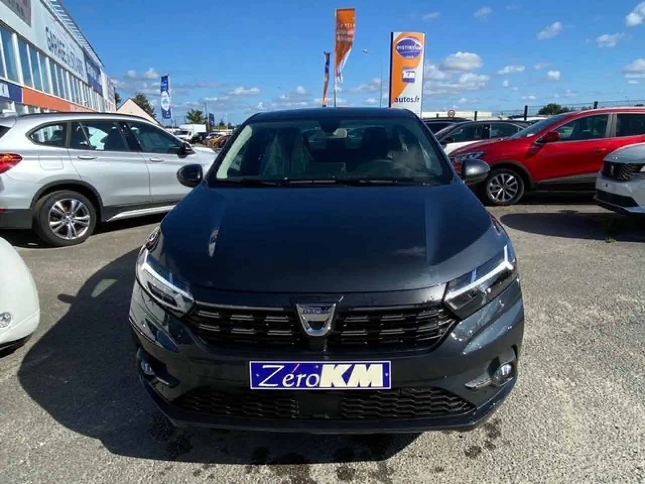 Vue de face d'une Dacia Logan gris foncé avec calandre noire et plaque "Zero KM" sur un parking extérieur.