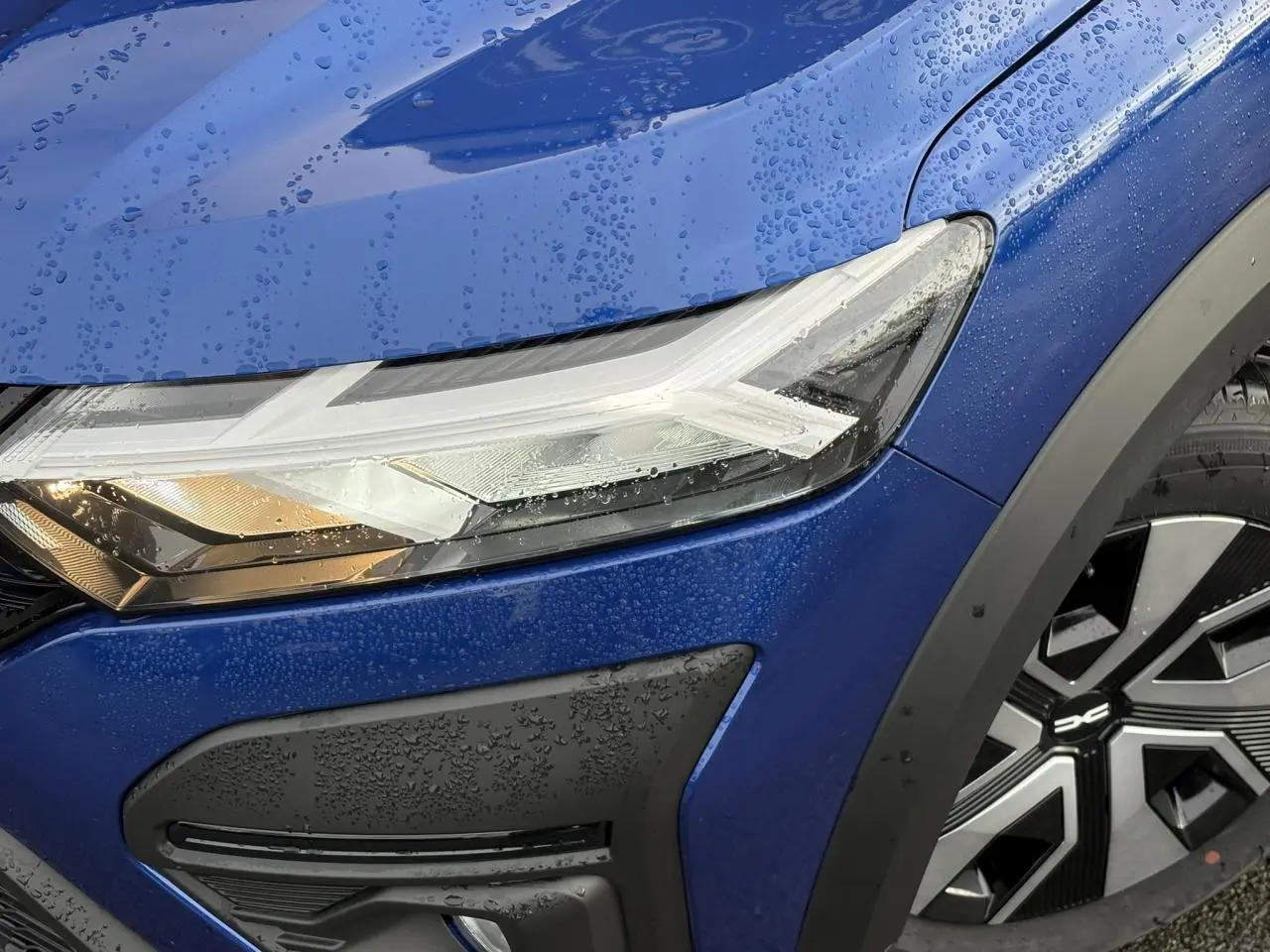 Gros plan sur le phare avant gauche et la roue avec enjoliveur 16" d'une Dacia Sandero Stepway Bleu Iron mouillée.