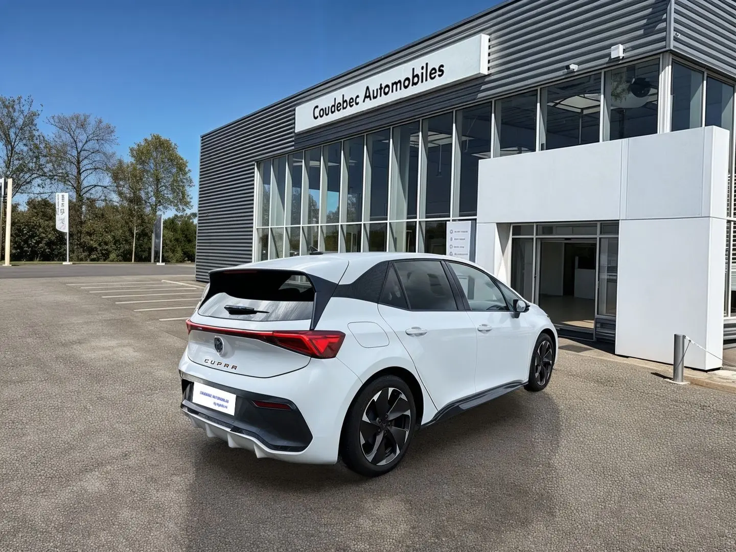 Vue 3/4 arrière droite d'une CUPRA Born blanche avec jantes noires Typhoon devant un bâtiment moderne.