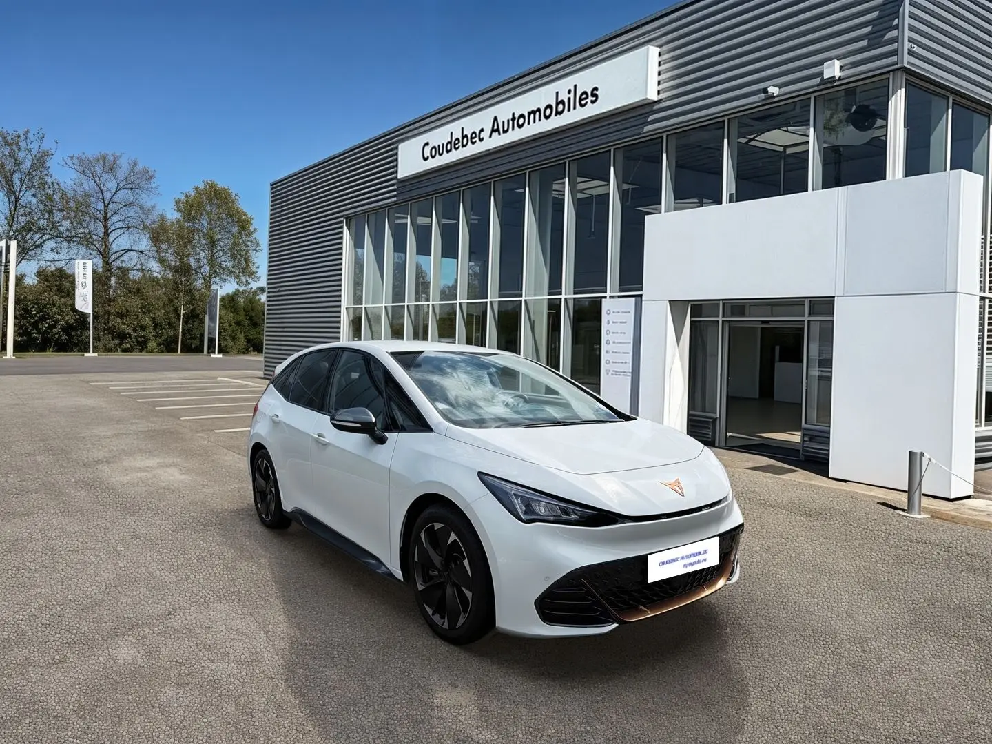 Vue 3/4 avant droite d'une CUPRA Born blanche avec jantes alliage noires devant un bâtiment de concession automobile.
