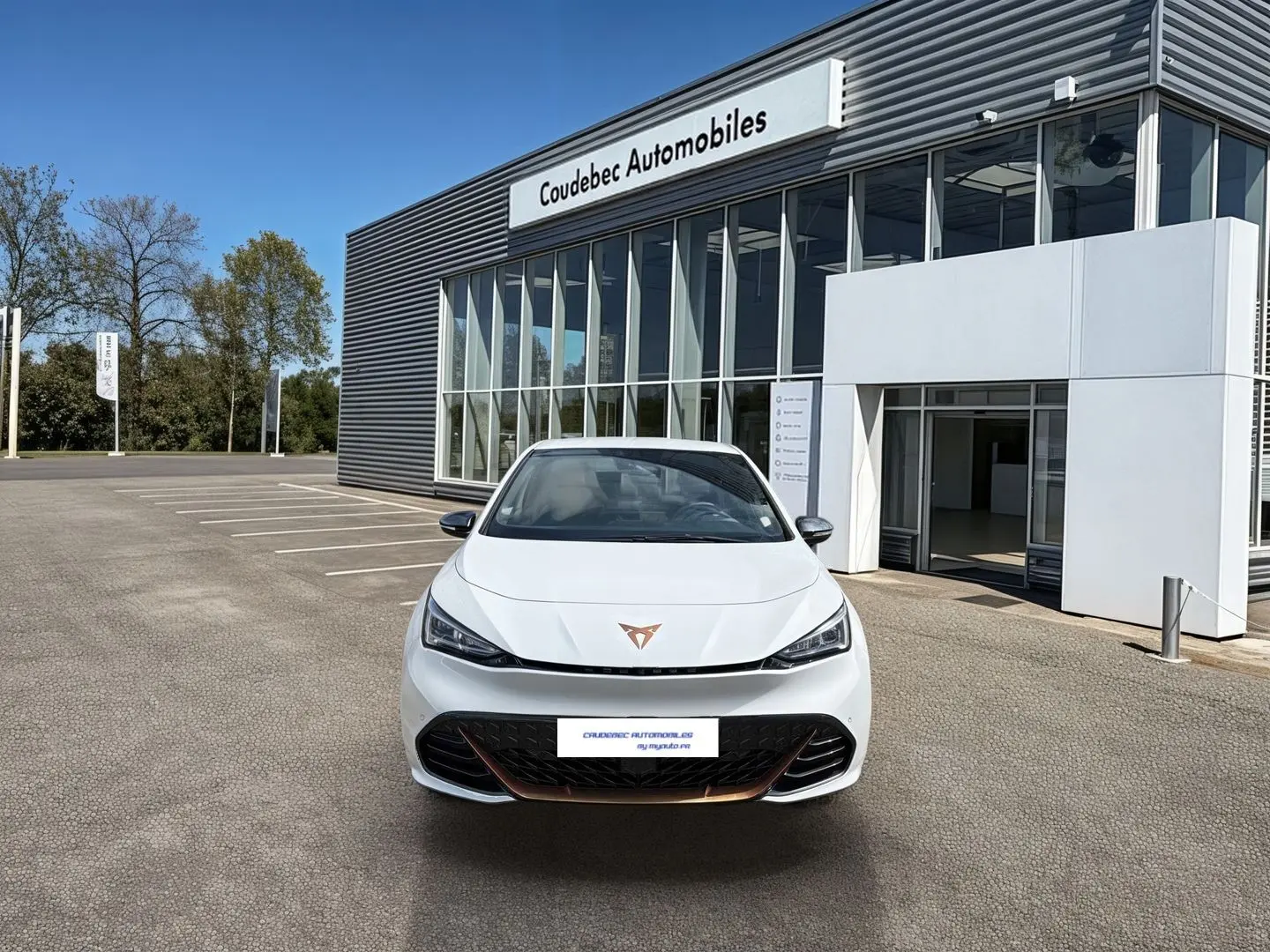 Vue avant d'une CUPRA Born électrique blanche avec logo cuivré, feux LED et calandre noire devant un bâtiment moderne.