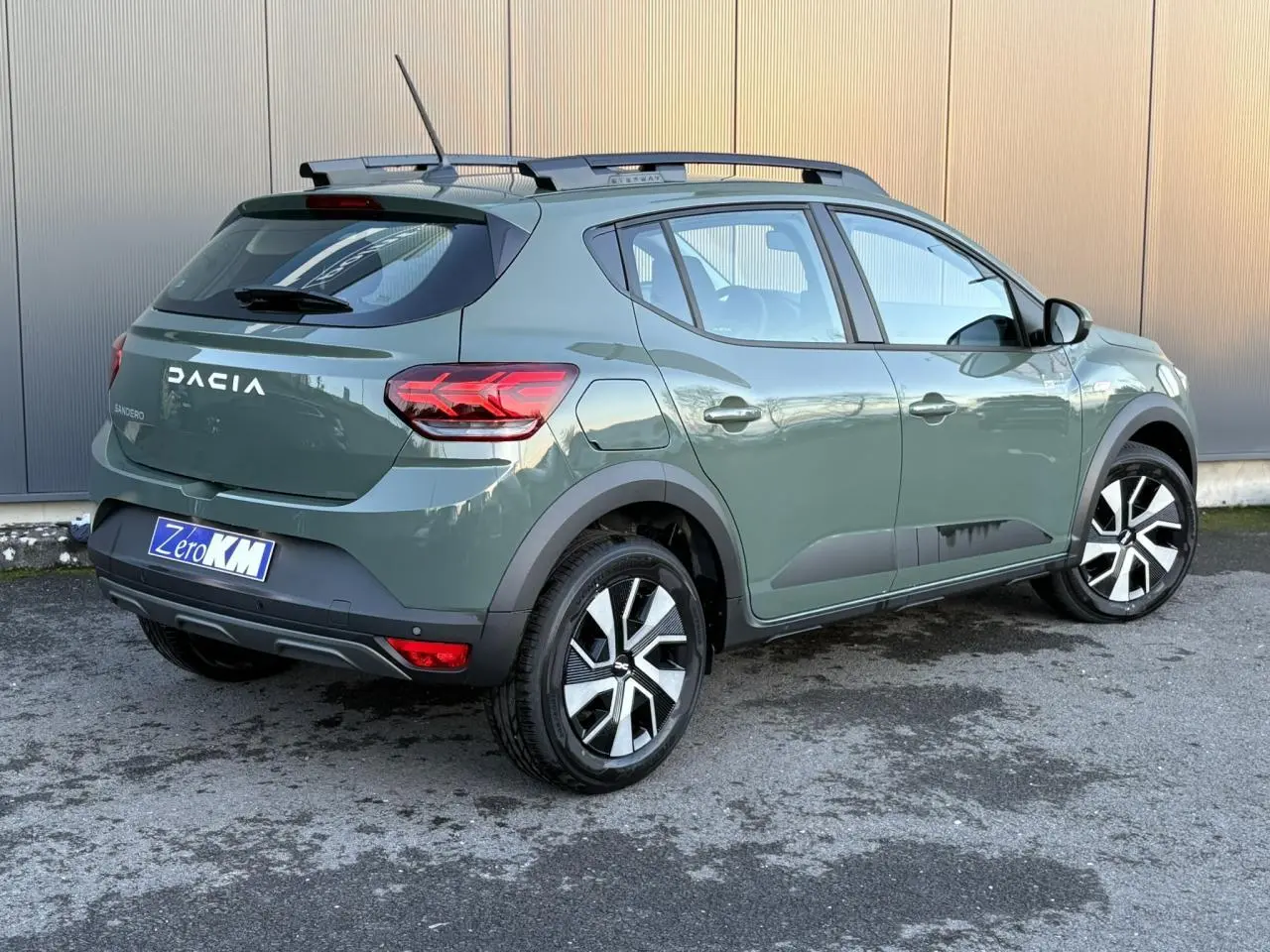 Vue 3/4 arrière droite d'une Dacia Sandero Stepway 2025 en kaki lichen avec barres de toit et jantes noires et argentées.