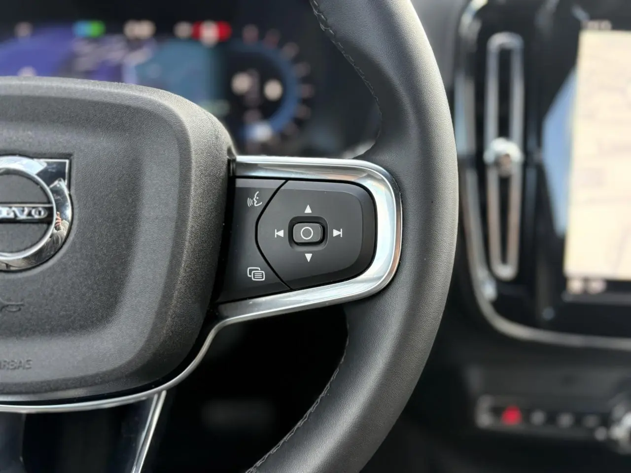 Gros plan sur le côté droit du volant noir d'un Volvo XC40 2024 avec commandes intégrées et tableau de bord flou.
