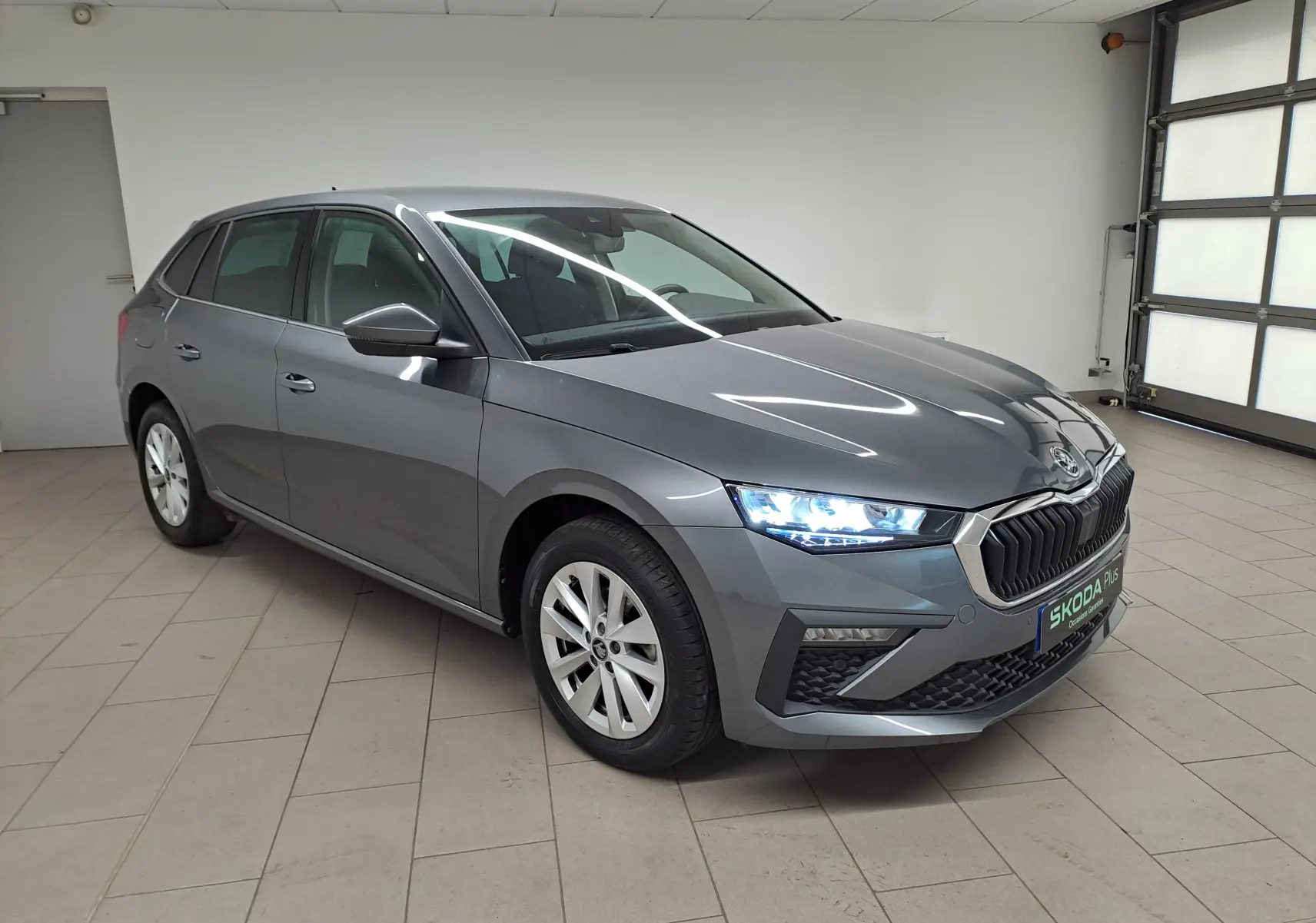 SKODA SCALA gris graphite en vue 3/4 avant droit avec phares allumés dans un garage lumineux.