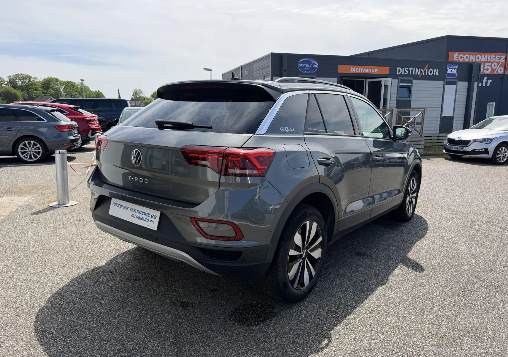 Vue 3/4 arrière droite d'un Volkswagen T-Roc gris avec barres de toit noires, stationné en extérieur devant un bâtiment commercial.