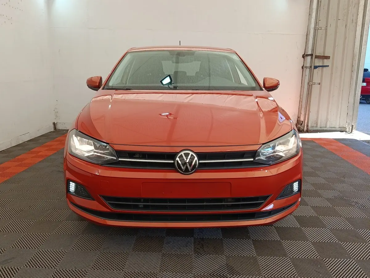 Vue de face d'une Volkswagen Polo 1.0 TSI 95 United orange avec phares et feux diurnes allumés dans un garage.