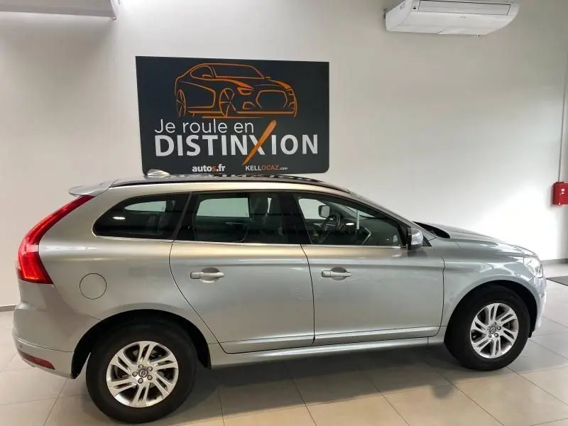 Profil côté gauche d'un Volvo XC60 argenté 2015 en intérieur, avec barres de toit et jantes alliage visibles.