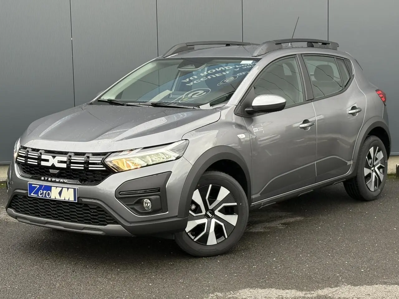 Vue 3/4 avant d'une Dacia Sandero Stepway 2025 gris schiste avec barres de toit et jantes spécifiques Atara Dark.