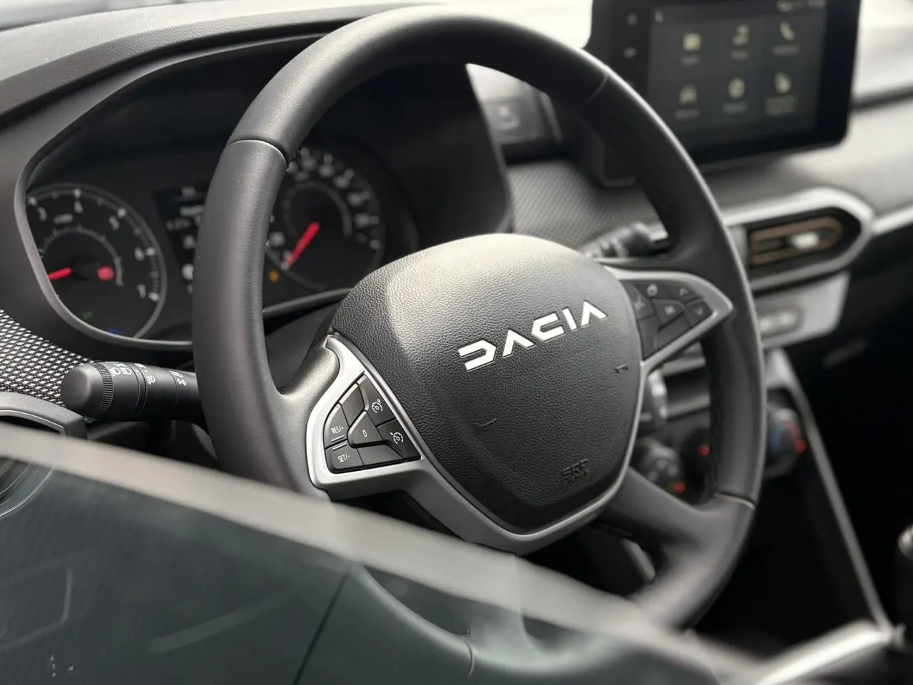 Vue rapprochée du volant noir Dacia avec commandes intégrées et tableau de bord numérique en arrière-plan.