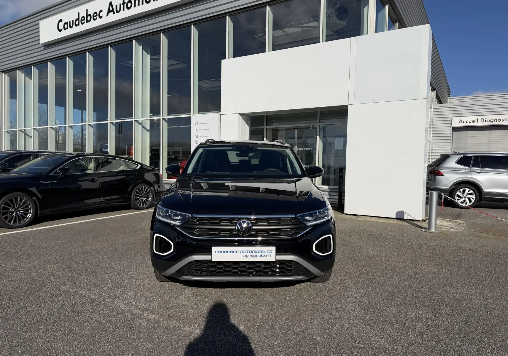 Volkswagen T-Roc noir vue de face, avec feux LED et calandre distinctive, devant un showroom Caudebec Automobiles.