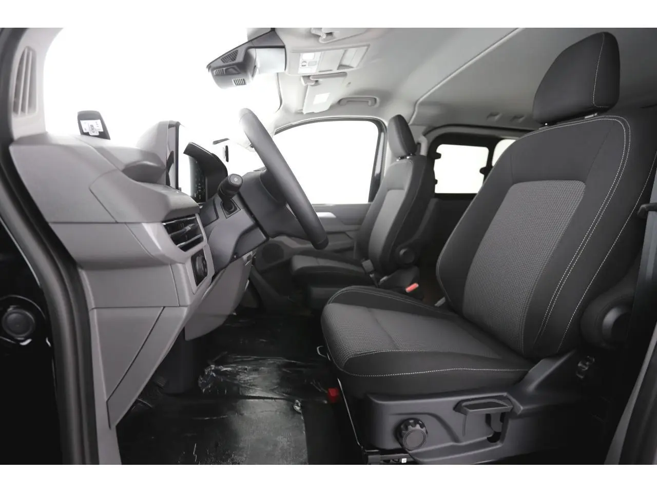 Vue intérieure côté conducteur du Volkswagen Transporter T7 2025, sièges tissu noir à rayures avec volant multifonction.