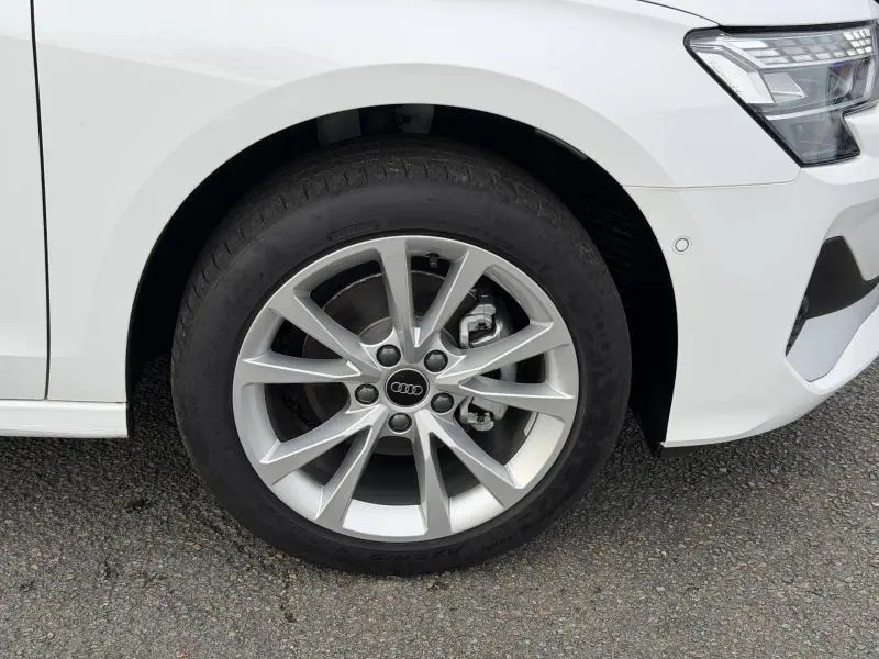 Gros plan sur la roue avant droite d'une Audi A3 Sportback blanche Arkona, avec jante alu et partie du phare avant visible.