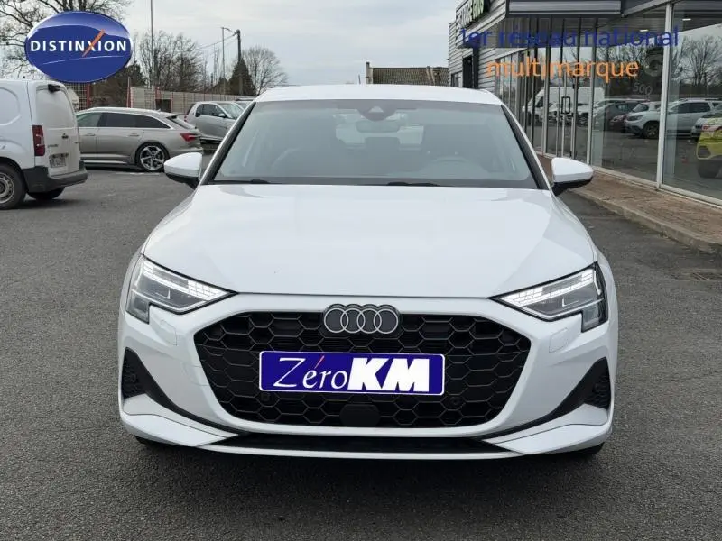 Vue de face d'une Audi A3 Sportback blanche Arkona avec calandre noire hexagonale et phares LED allumés.