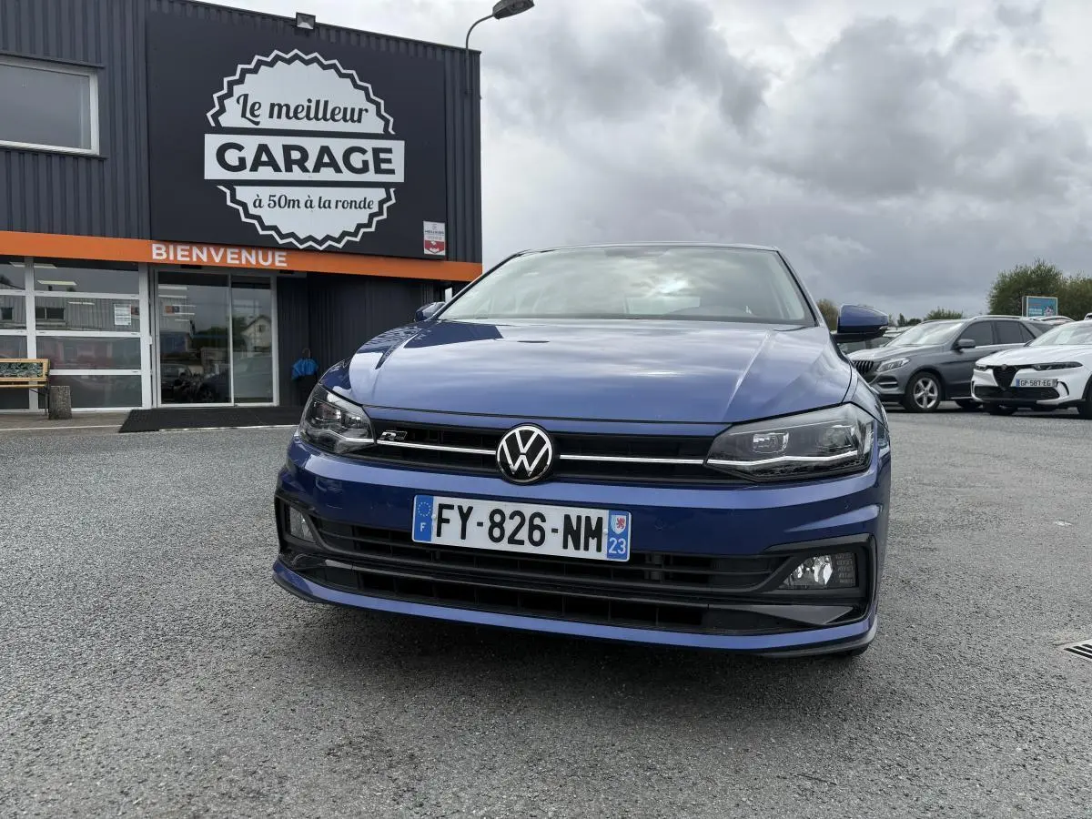 Vue de face d'une Volkswagen Polo 1.0 TSI R-Line bleu métallisé avec calandre et badge R visibles, stationnée devant un garage.