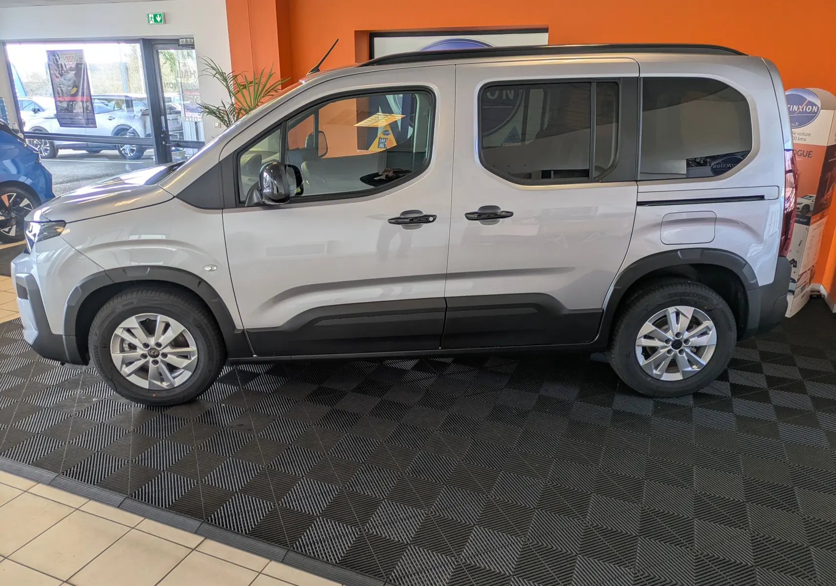 Vue de profil côté gauche d’un Peugeot Rifter gris Artense 2025 avec barres de toit noir brillant en intérieur showroom.