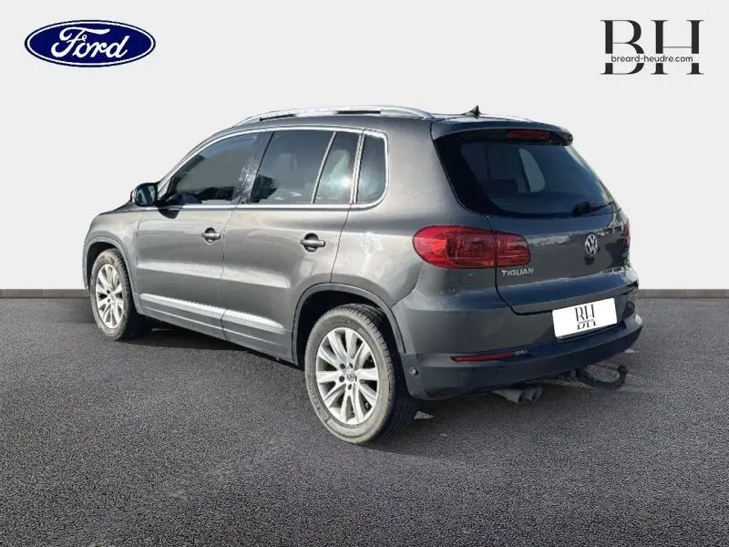 Vue 3/4 arrière droite d'un Volkswagen Tiguan gris Crypton avec barres de toit et attelage visible.