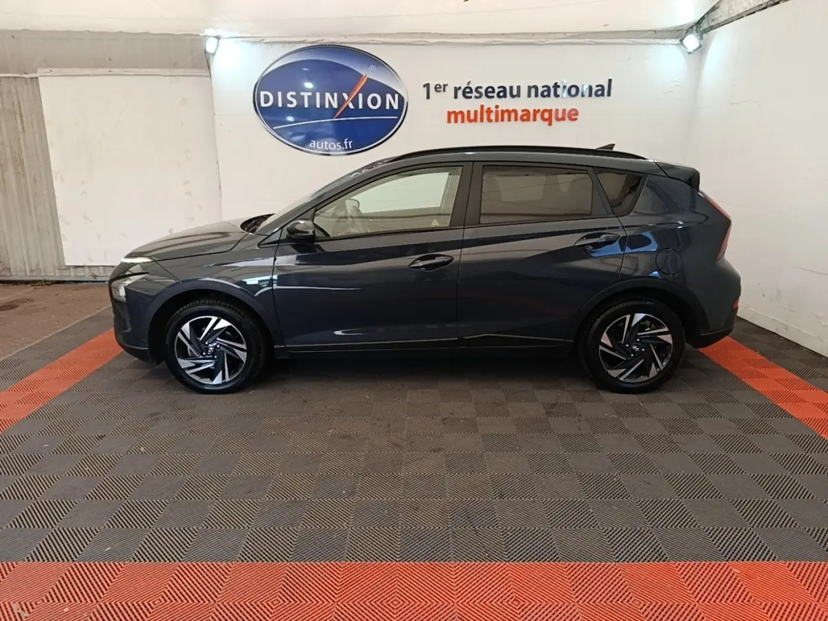 Profil côté gauche d'une Hyundai Bayon gris foncé 2023, avec jantes alliage et toit noir en arrière-plan intérieur showroom.
