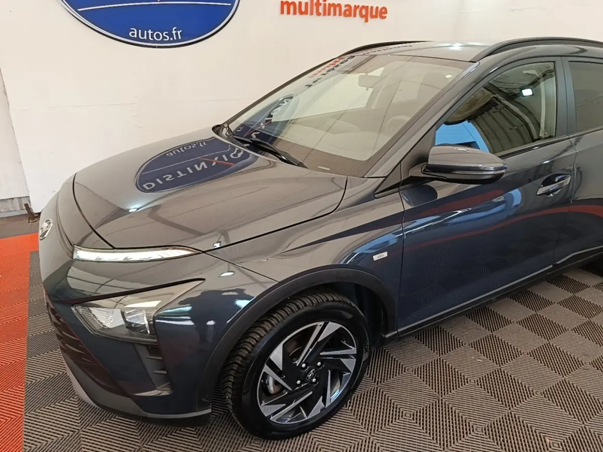Vue 3/4 avant gauche d'une Hyundai Bayon gris foncé avec jantes alliage et badge hybride visible sur l'aile avant.