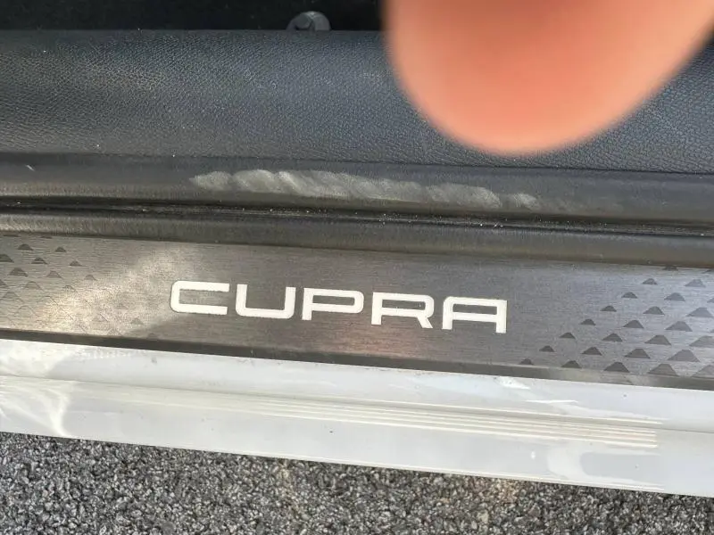 Gros plan sur la plaque de seuil de porte CUPRA noire avec logo blanc, sur un véhicule noir métallisé, vue côté gauche.