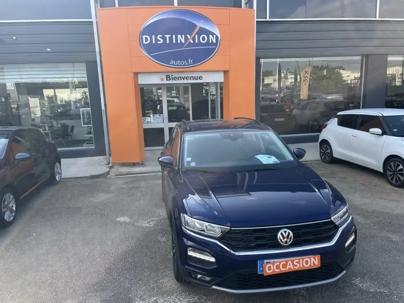 Volkswagen T-Roc bleu foncé en 3/4 avant garé devant un concessionnaire avec panneau orange "Distinxion".