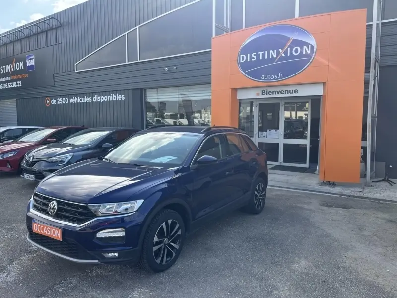 Volkswagen T-Roc gris en 3/4 avant droit, stationné devant un concessionnaire Distinxion avec panneau orange.