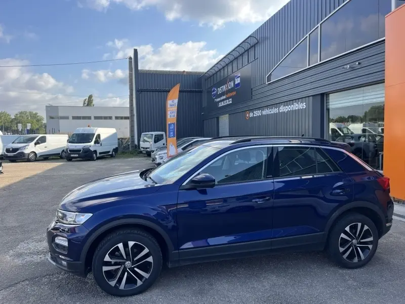 Vue latérale droite du Volkswagen T-Roc bleu foncé garé devant un bâtiment commercial avec jantes alliage noires et argent.