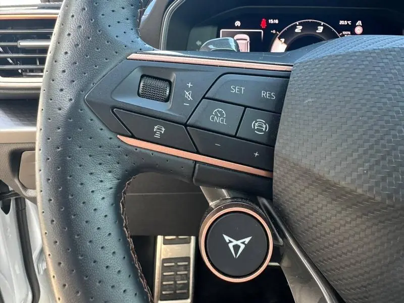 Gros plan sur le volant perforé noir avec commandes intégrées et logo CUPRA, tableau de bord numérique en arrière-plan.