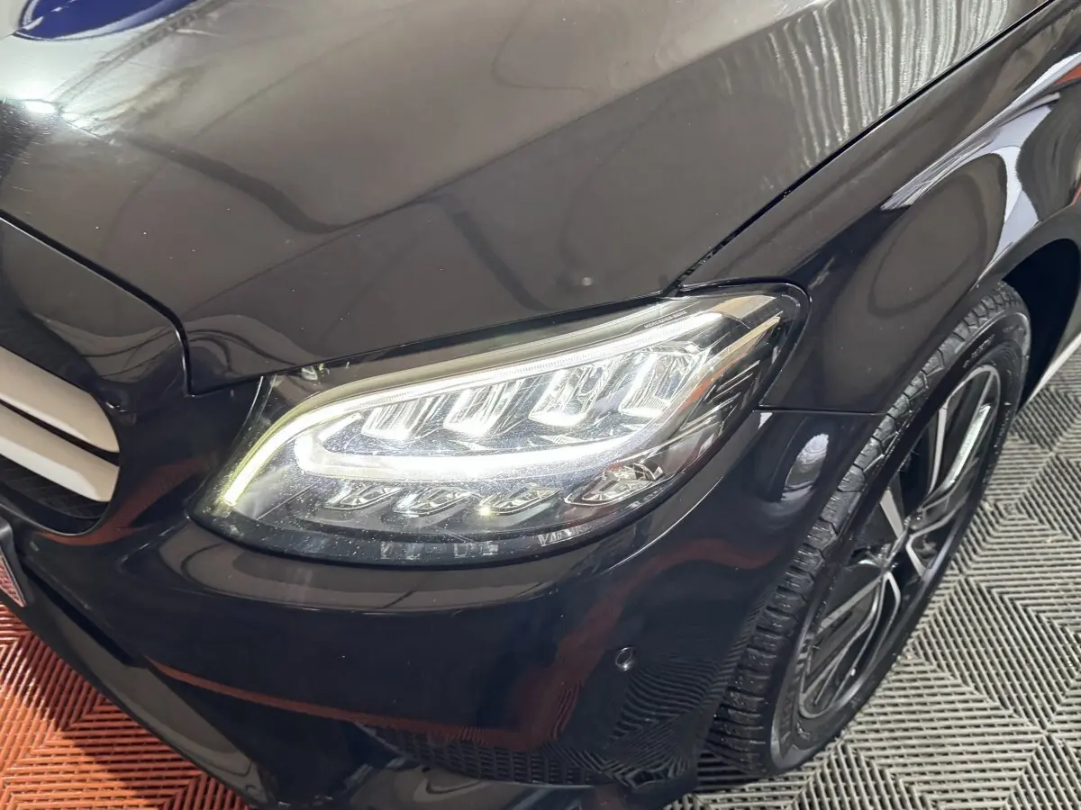 Gros plan sur le phare avant droit allumé d'une Mercedes Classe C Break noire, avec jante alliage visible.
