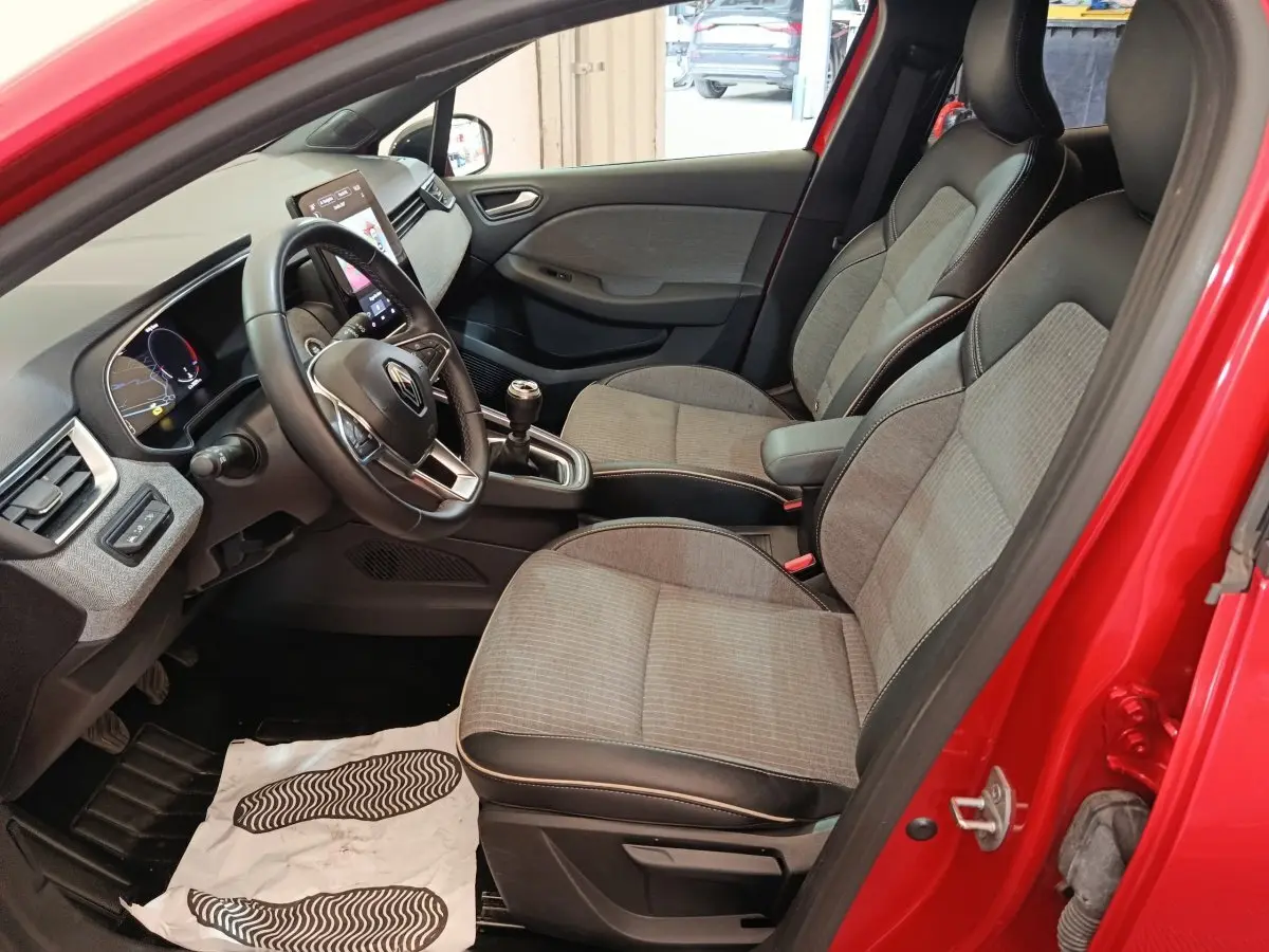 Vue intérieure côté conducteur d'une Renault Clio rouge 2024, sièges tissu gris avec accoudoir central et volant multifonctions.