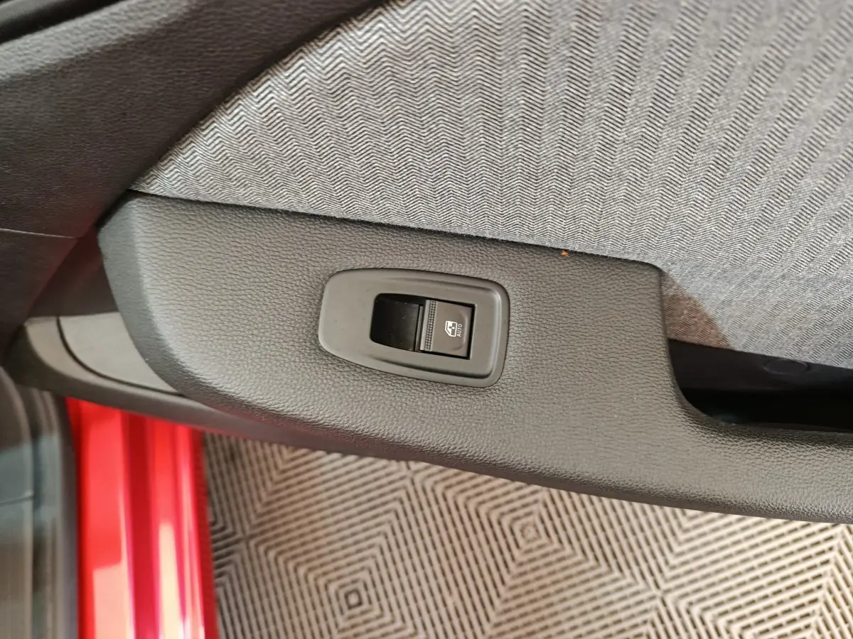 Gros plan sur le bouton de commande de lève-vitre côté conducteur, avec partie de la porte rouge visible.