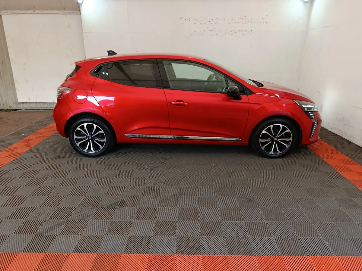 Profil droit d'une Renault Clio rouge 2024 avec jantes alliage et feux LED distinctifs, sur sol damier gris et orange.