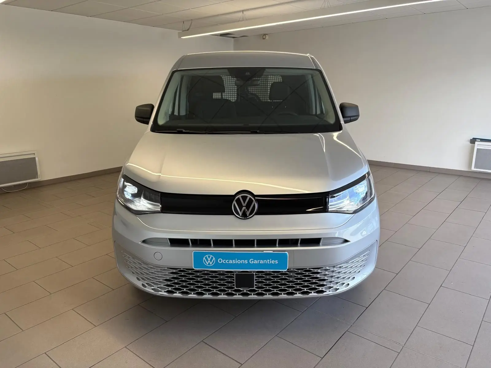 Vue frontale d'un Volkswagen Caddy 2025 gris argent avec calandre noire et plaque "Occasions Garanties" en intérieur.
