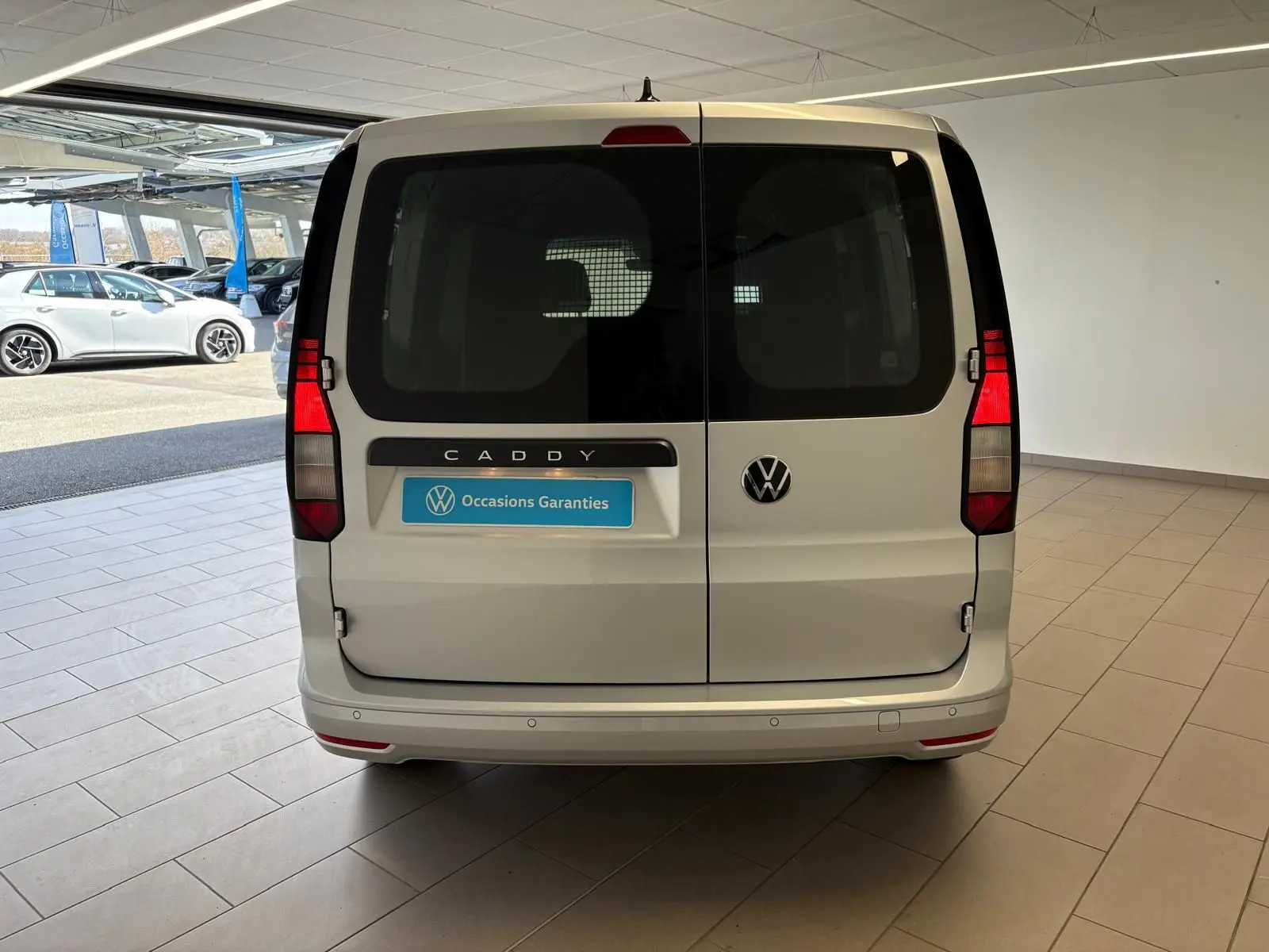 Vue arrière d'un Volkswagen Caddy gris argent 2025 avec portes battantes et logo VW visible en concession.