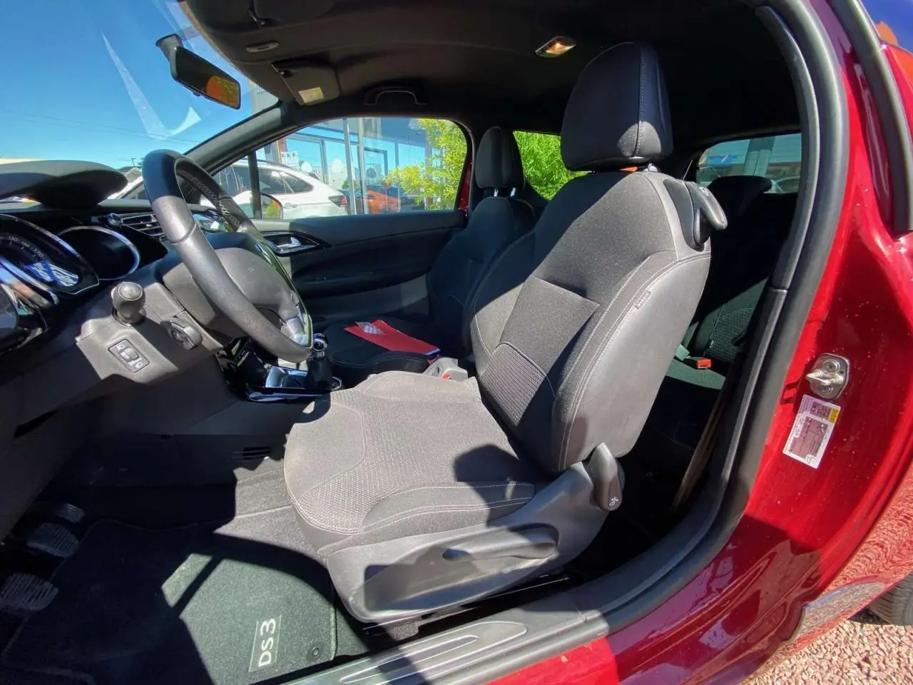 Vue intérieure côté conducteur d'une DS3 rouge avec sièges tissu gris et tableau de bord noir lumineux.