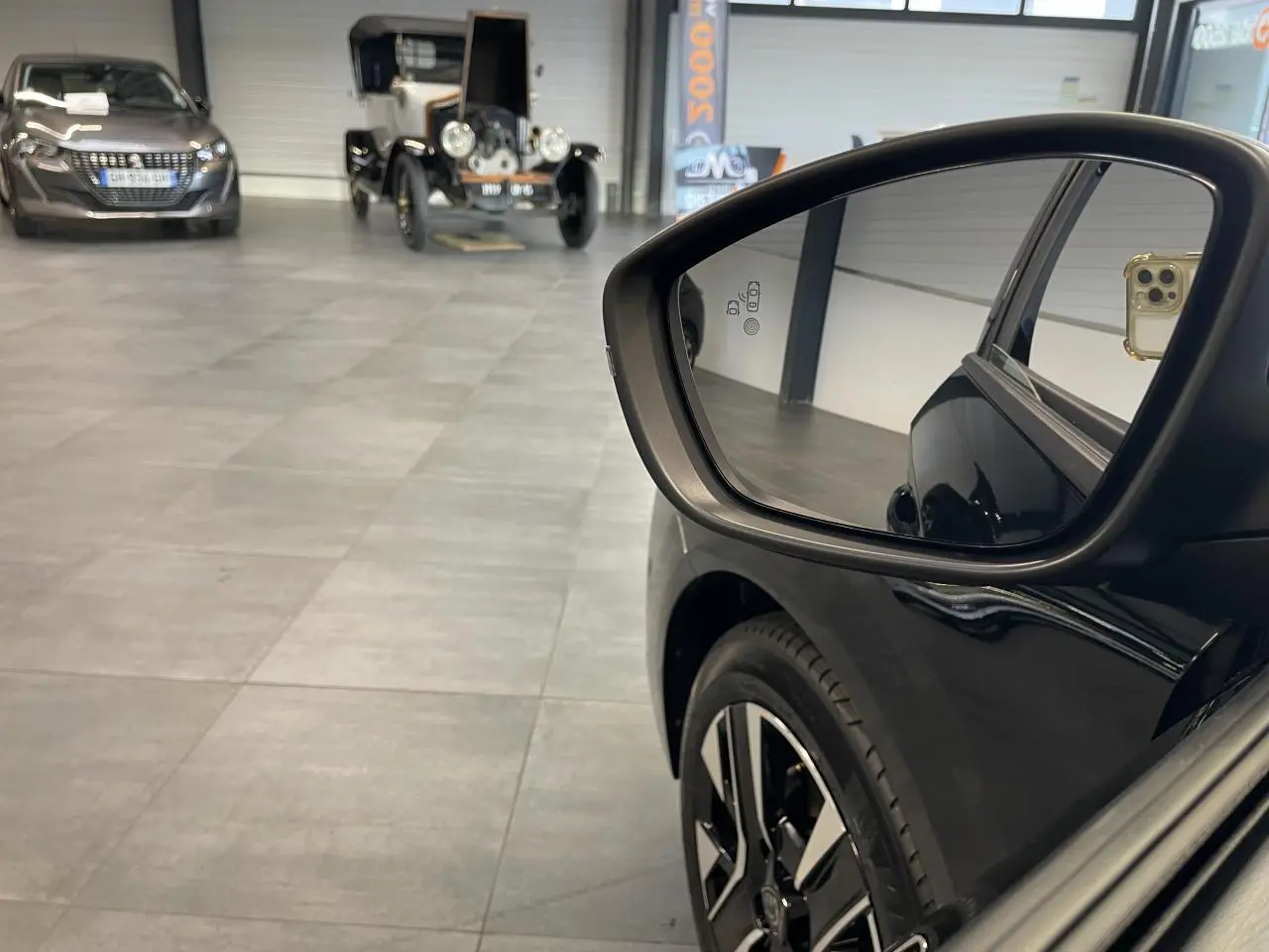Rétroviseur côté droit d'une Peugeot 208 noire 2025 avec jante alliage bicolore et reflet intérieur showroom.
