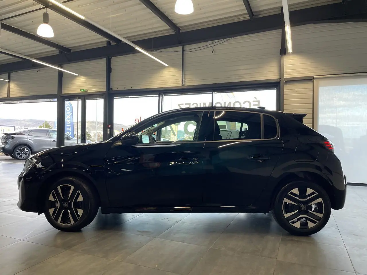Profil latéral droit d'une Peugeot 208 noire métallisée 2025 en showroom avec jantes alliage bi-ton et feux arrière à LED.