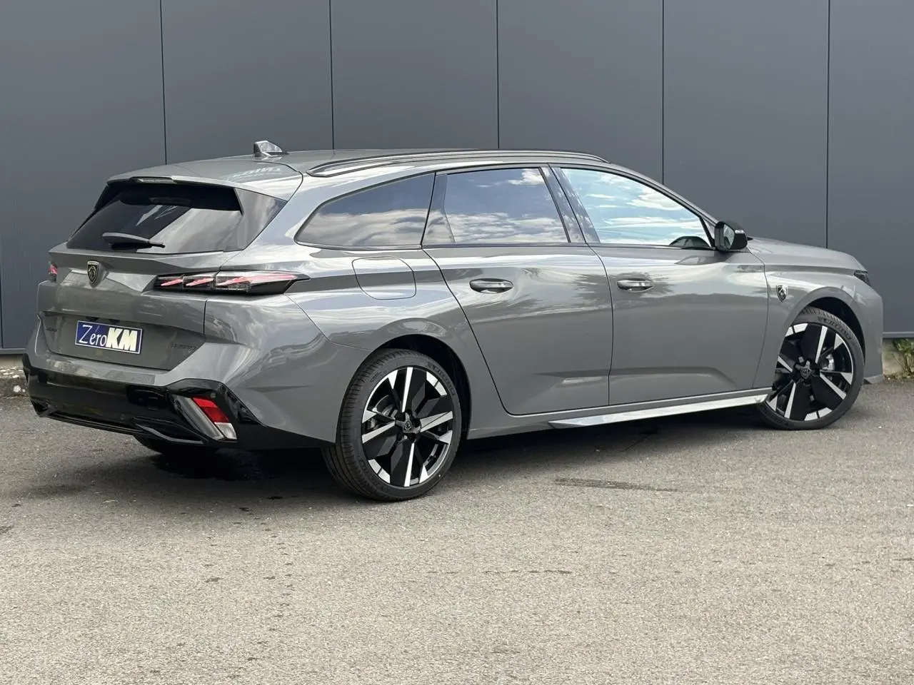 Vue 3/4 arrière droite d'une Peugeot 308 SW gris Sélénium avec jantes bi-ton et barres de toit noires.