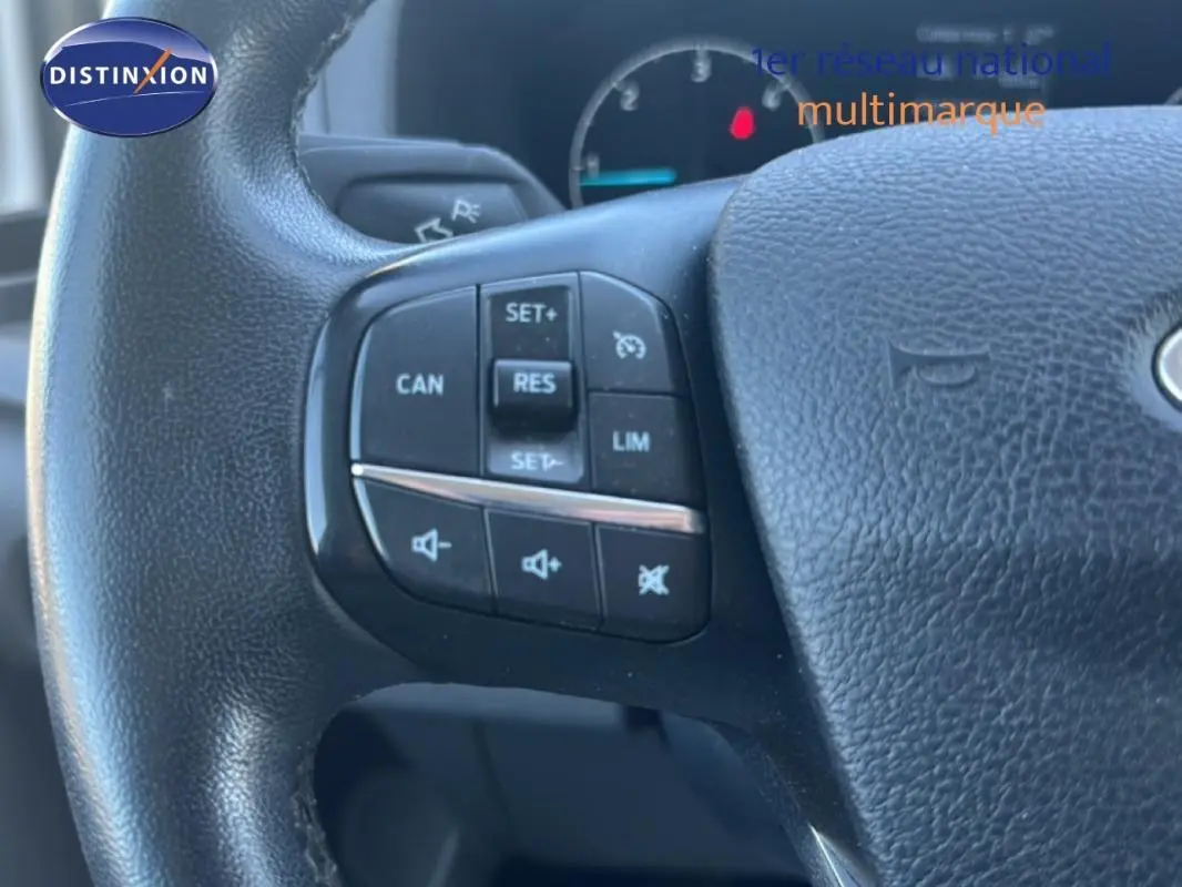 Gros plan sur les commandes du volant cuir noir du Ford Transit Custom Fourgon blanc, avec régulateur de vitesse et contrôle audio.
