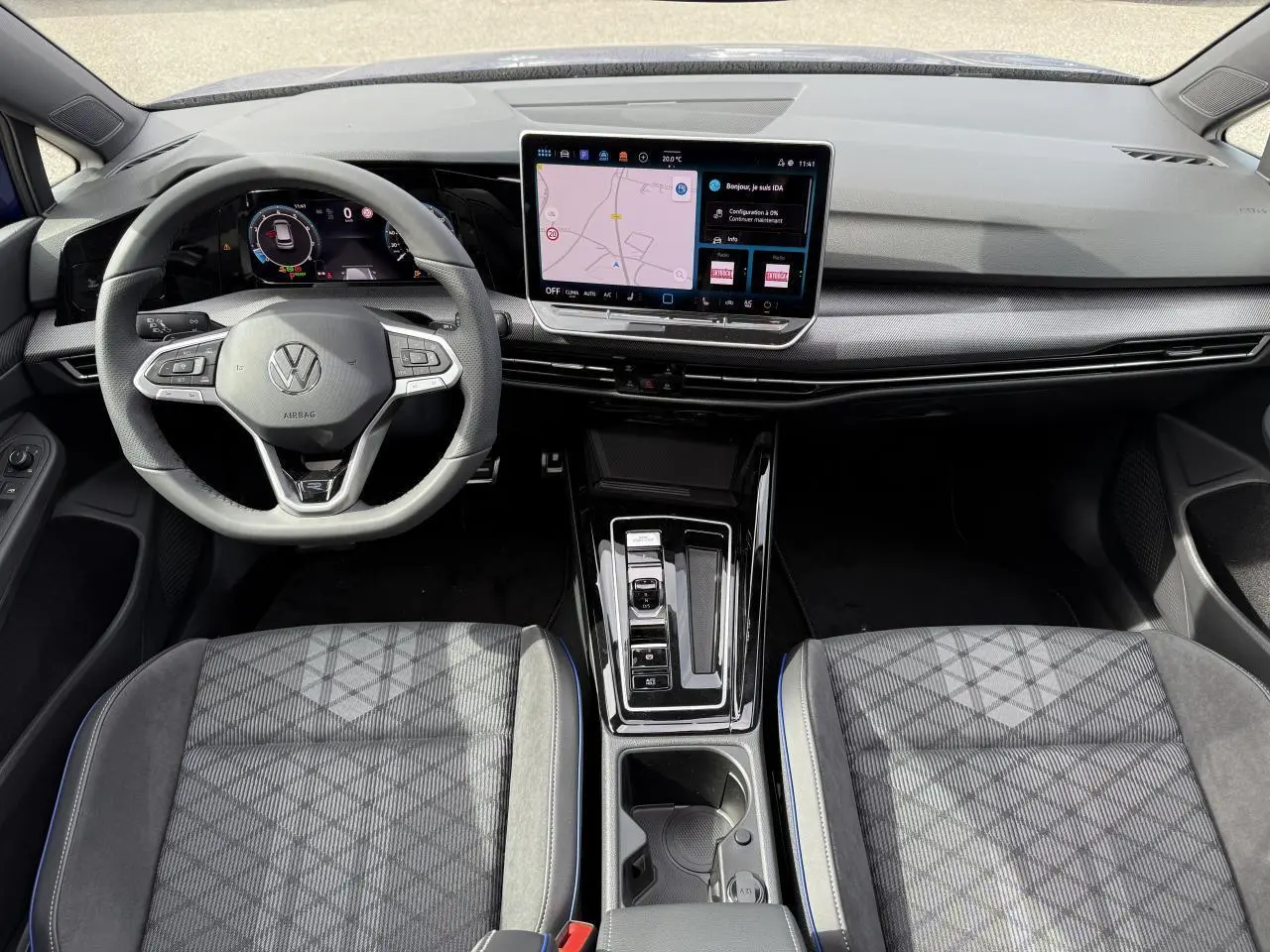 Vue intérieure avant de la Volkswagen Golf VIII R-Line 2025, tableau de bord noir avec écran tactile central et sièges tissu gris à motifs.