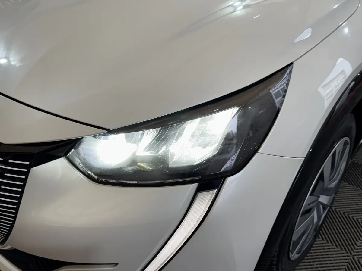 Gros plan sur le phare avant droit allumé d'une Peugeot 208 électrique blanche, avec partie de la roue visible.
