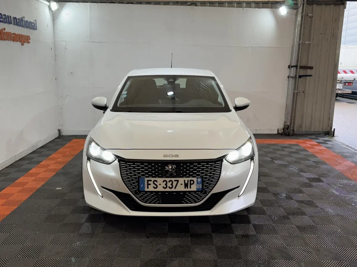 Peugeot 208 électrique blanche vue de face, avec phares LED allumés dans un garage.