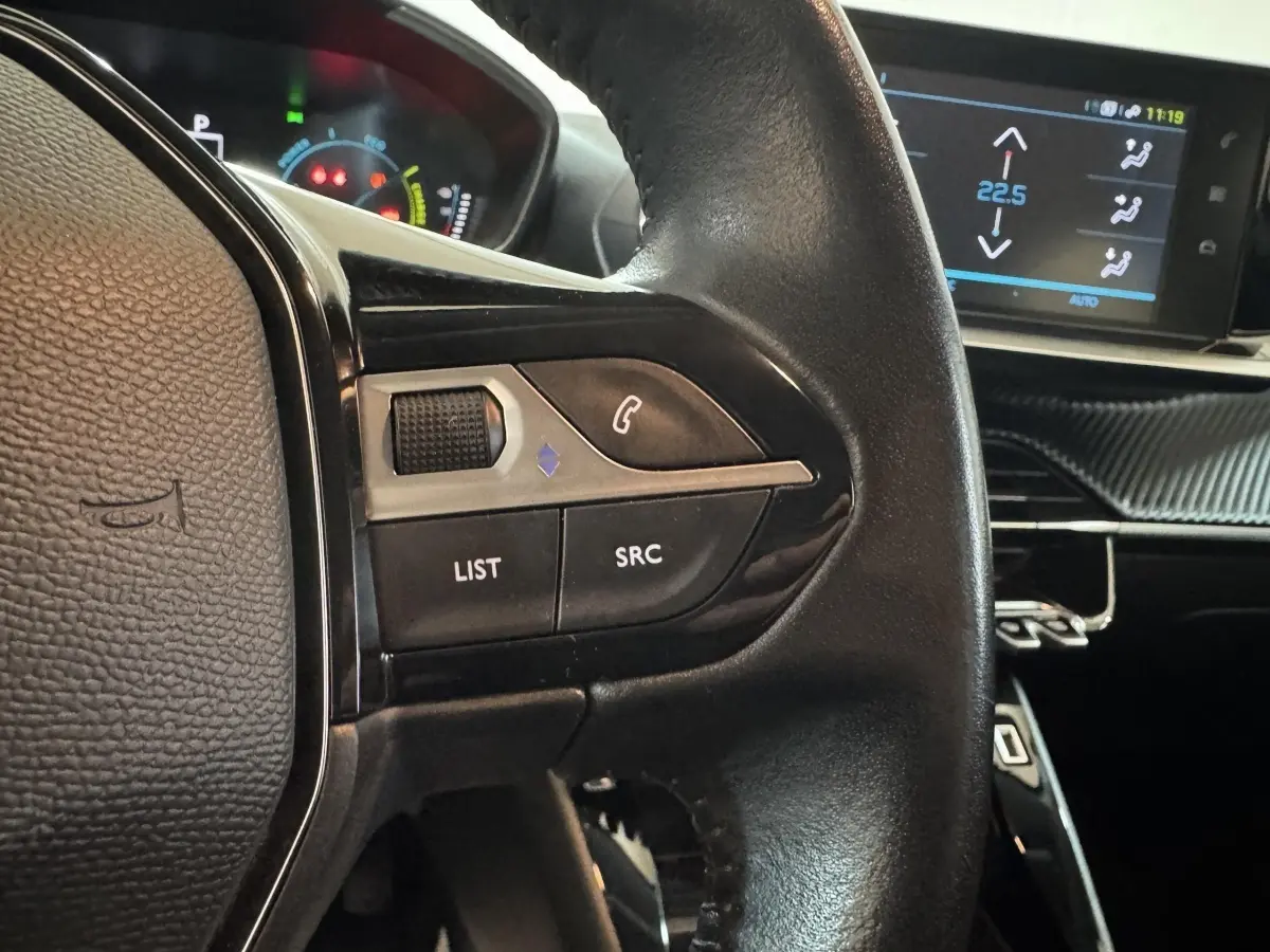 Gros plan sur les commandes gauche du volant cuir noir de la Peugeot 208 électrique Active Business avec écran tactile en arrière-plan.