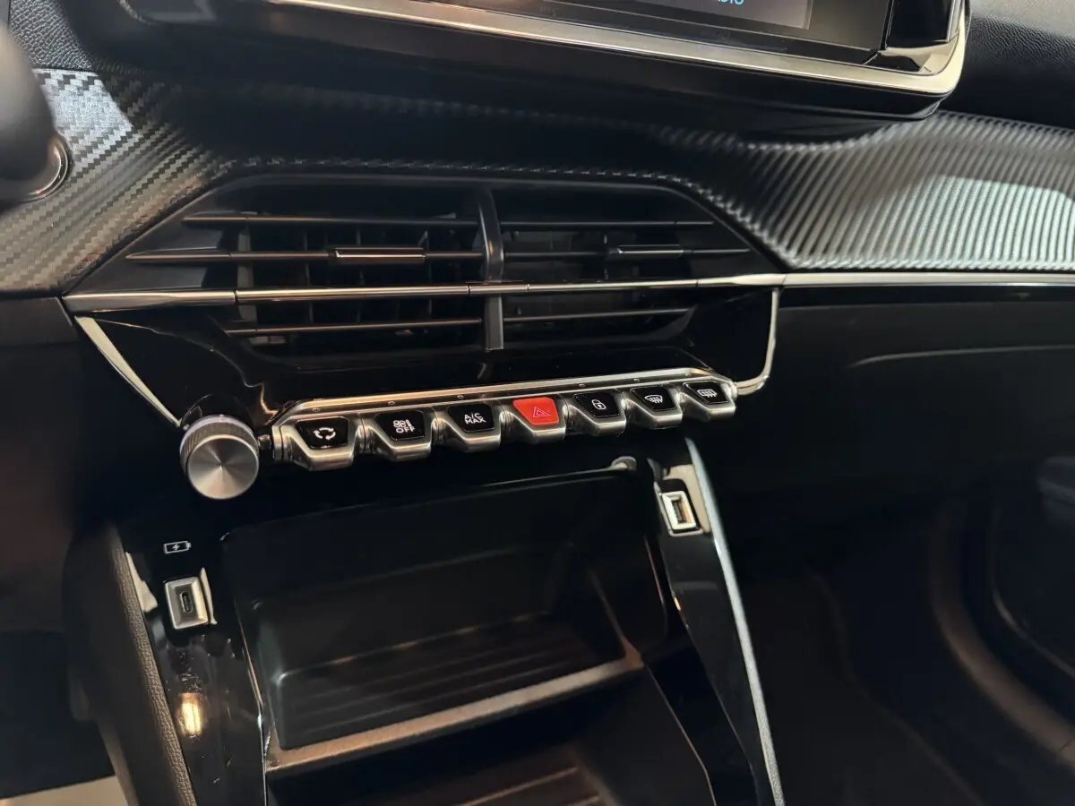 Gros plan sur la console centrale noire de la Peugeot 208 électrique, avec boutons métalliques et finition carbone.