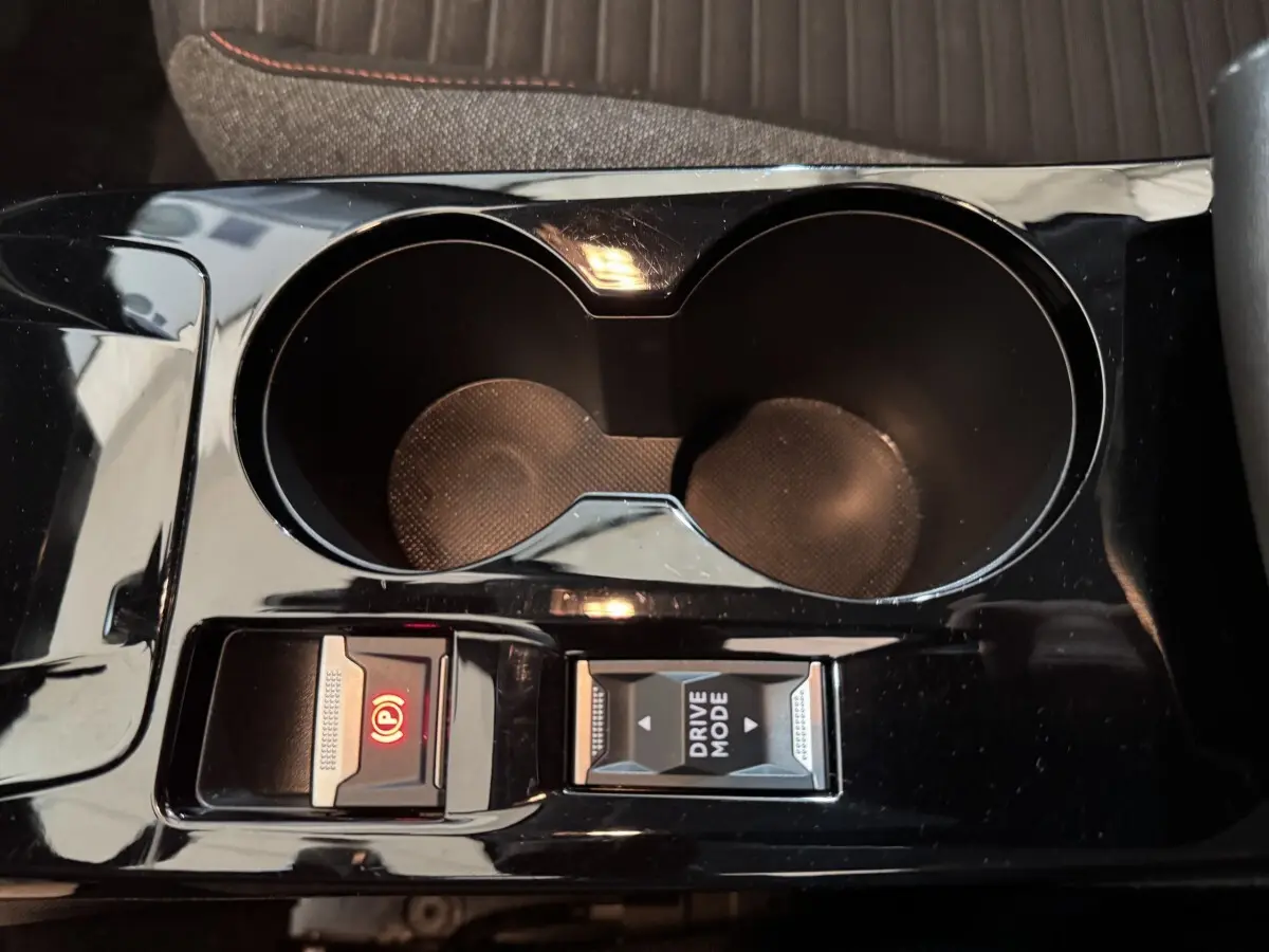 Gros plan sur la console centrale noire brillante de la Peugeot 208 électrique avec porte-gobelets et boutons frein à main et mode de conduite.