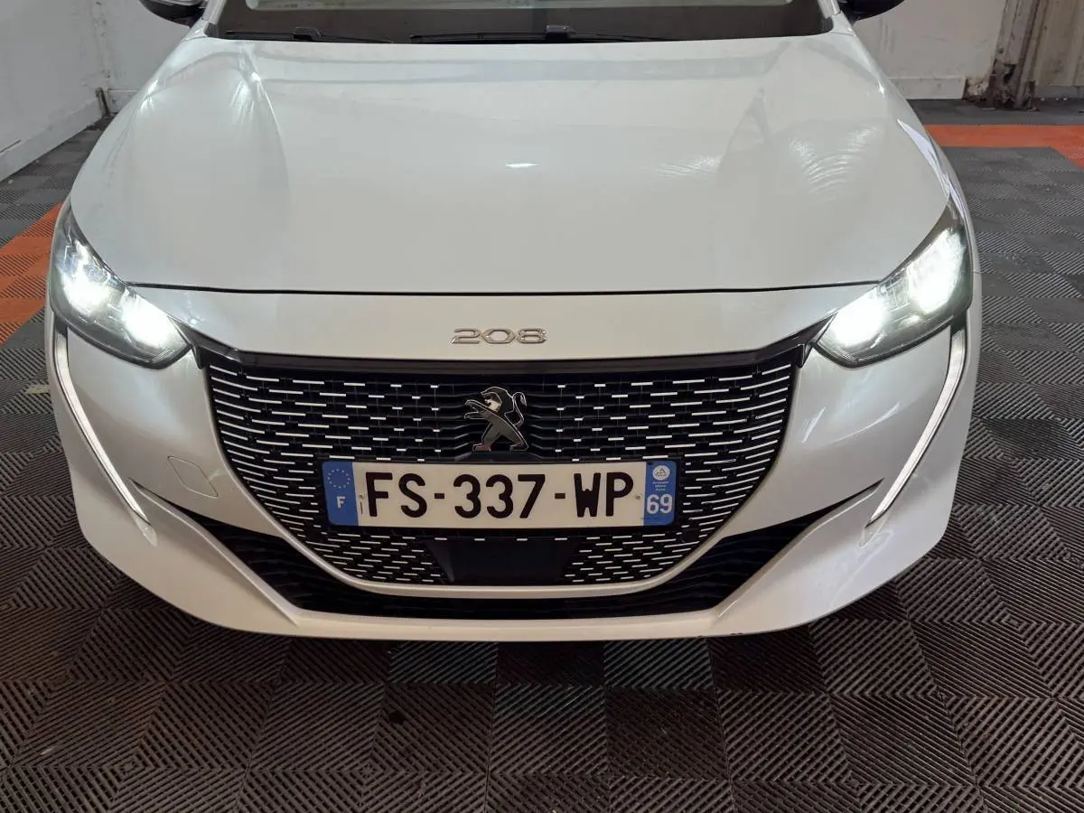 Vue avant rapprochée d'une Peugeot 208 électrique blanche avec phares LED allumés et calandre noire distinctive.